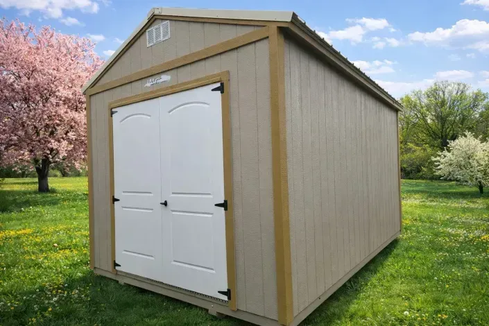 Beige garden shed with white double doors and a peaked roof on grass, with trees in the background
