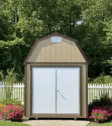 Small beige lofted storage shed with white double doors in a flowered yard, framed by a white picket fence and trees
