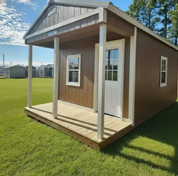 Small brown cabin with white trim and porch on a grassy lot under a blue sky