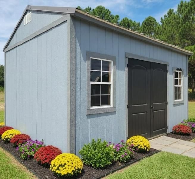Gray garden shed with black double doors, white-trim windows, and colorful flower beds