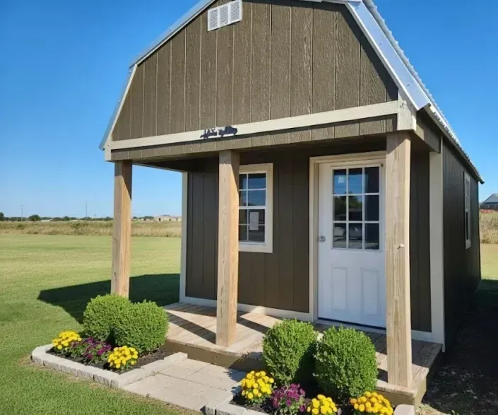 Small brown shed with white door and porch, set beside a grassy field with flowers in front.