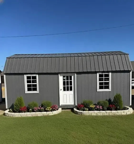 Small gray shed with white door and windows, black roof, and flower beds on a green lawn under a blue sky