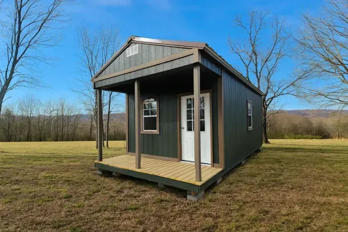 Small gray cabin with a front porch in a grassy field under a blue sky