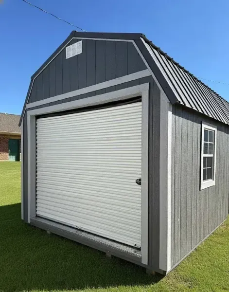 Small gray storage shed with a white roll-up door on a grassy lawn under a blue sky