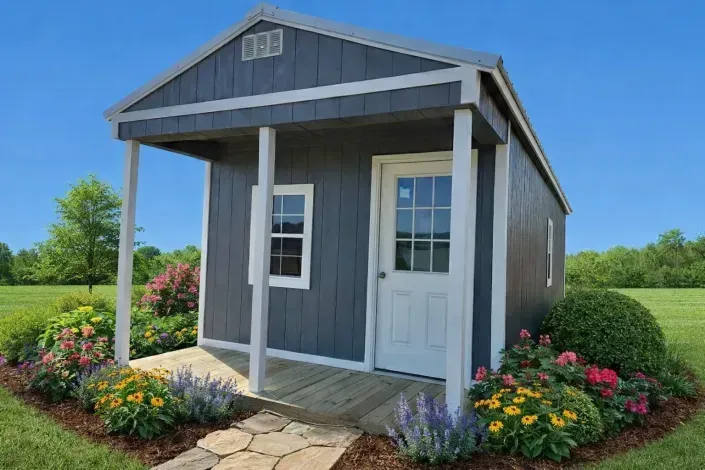 Small gray shed with white doors and porch, surrounded by flowers in a sunny garden.