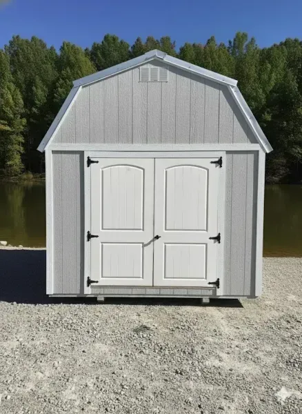 White and gray storage shed with double doors on a gravel lot by a lake and trees