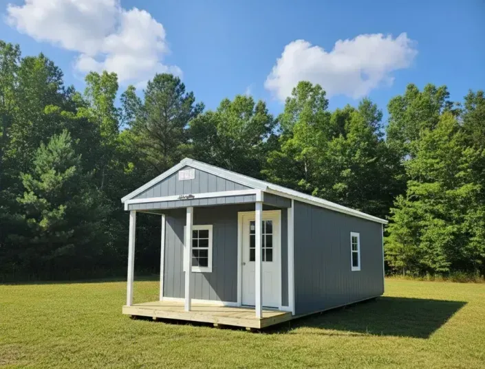 Small gray cabin with white trim and porch sitting in a grassy field near trees under a blue sky