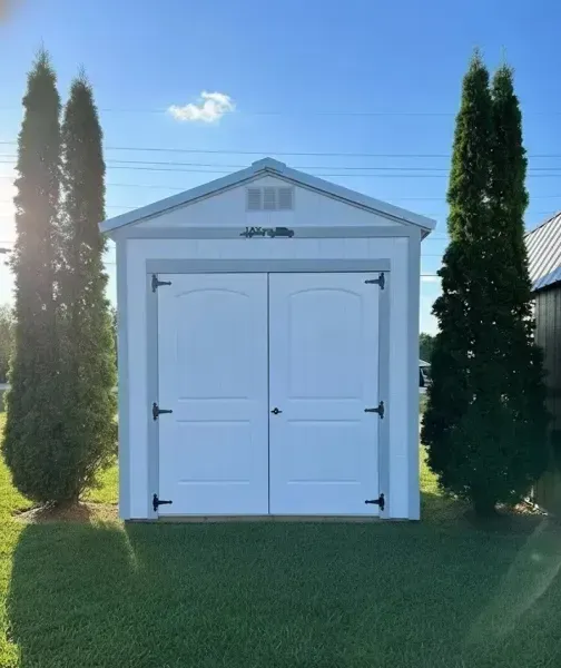 White garden shed with double doors, framed by two tall evergreen trees on a grassy lawn under a blue sky.