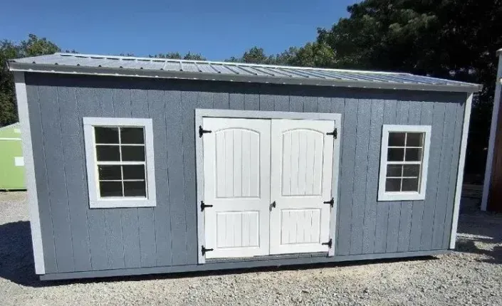 Small gray shed with white double doors and two windows on a gravel lot outdoors