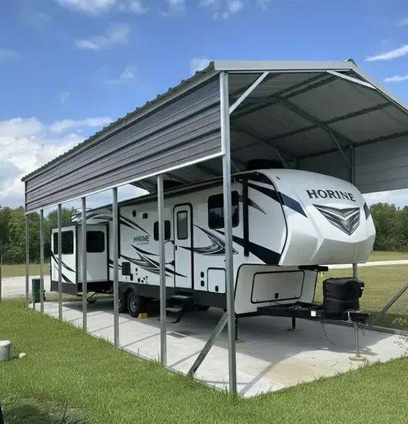 White travel trailer under a metal carport on a concrete pad, in a grassy field under a blue sky
