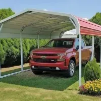Red pickup truck parked under a white carport beside a red-roofed shed in a grassy yard