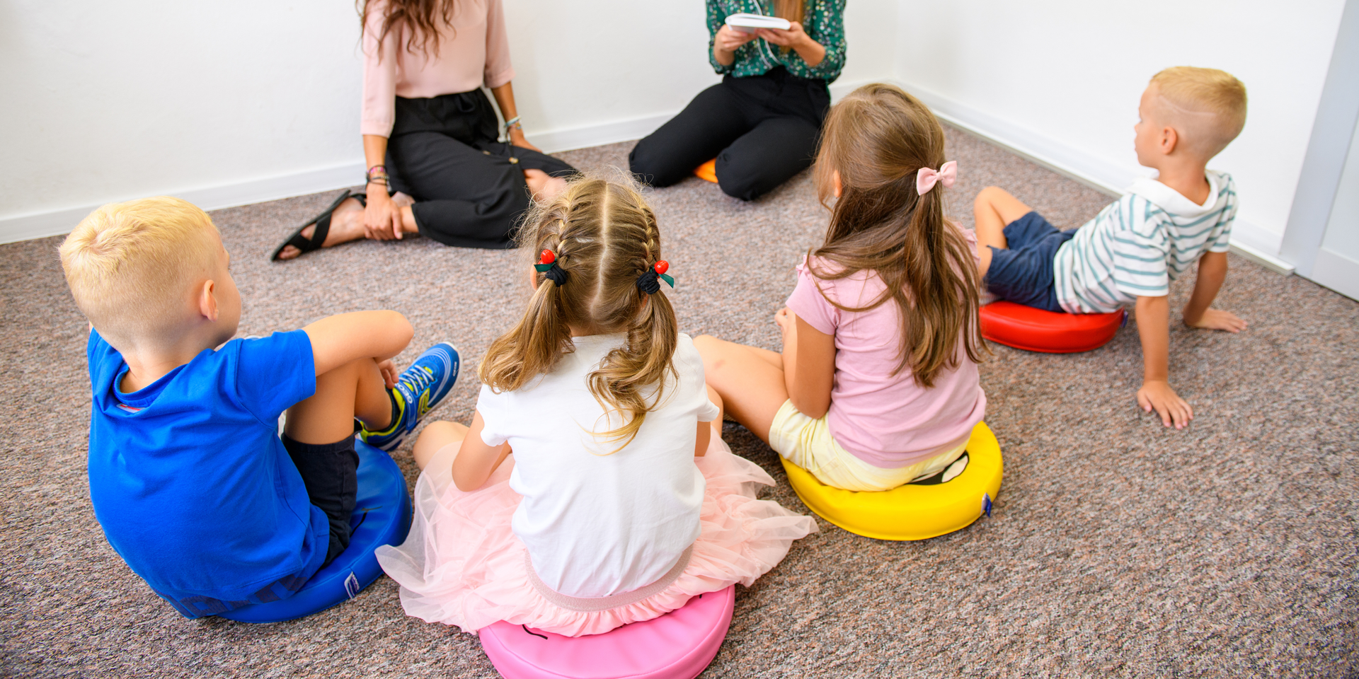 Children sitting on colorful cushions, in a circle, with two adults. Indoor setting on a carpeted floor.