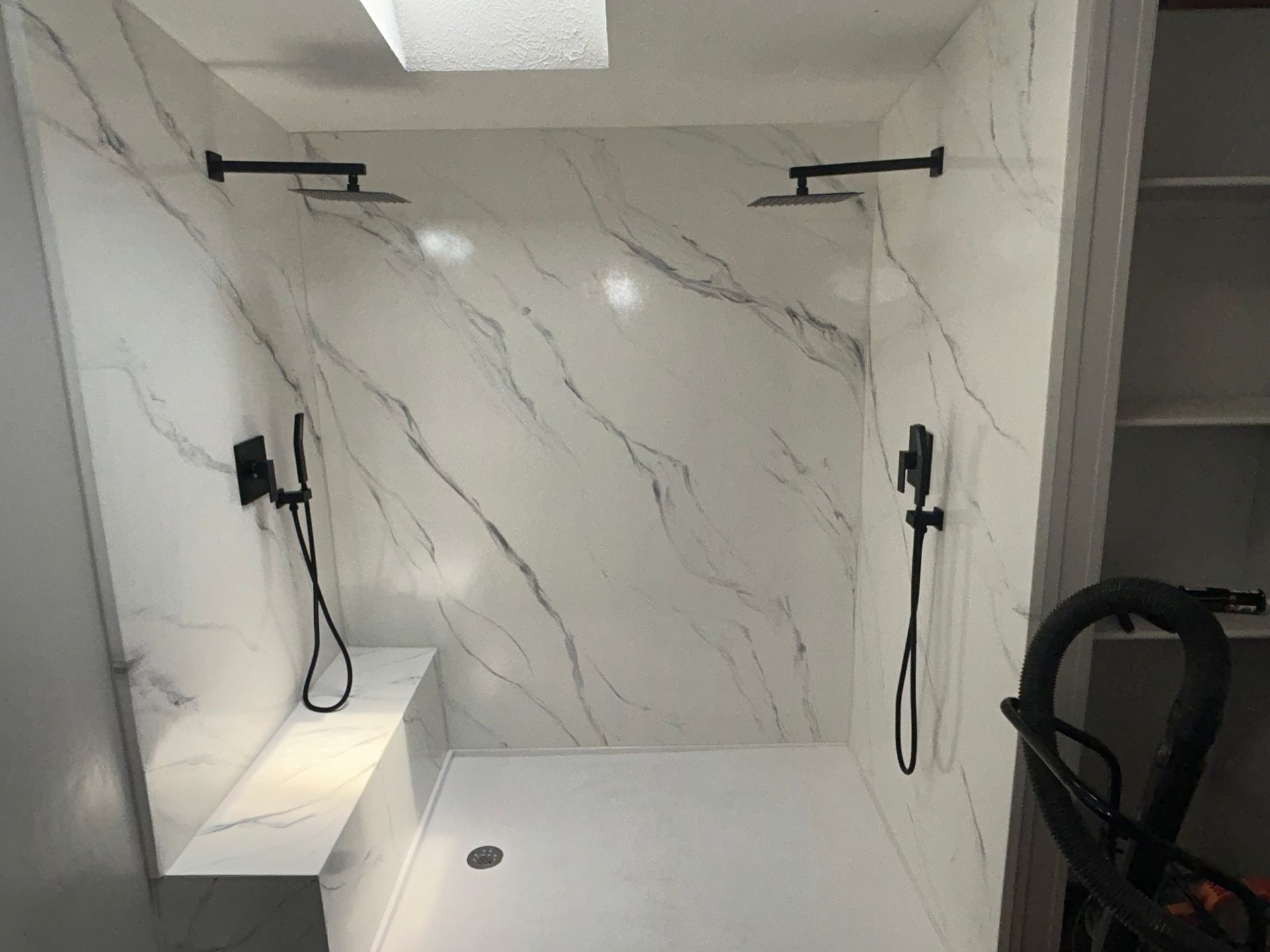 Modern shower with marble-look walls, two black showerheads, bench, and white floor.