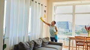 Woman wearing gloves, cleaning curtains in a bright room with a sofa, table, and window.