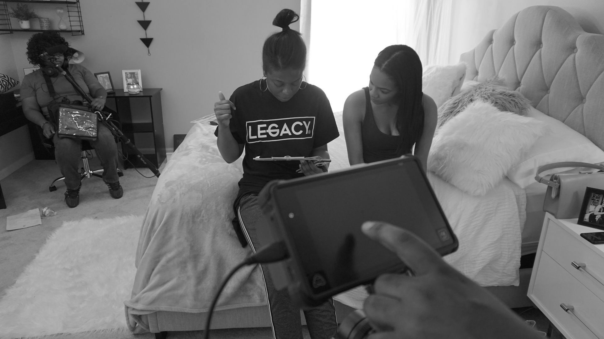 People seated on a bed in a bedroom while someone holds a tablet in the foreground.