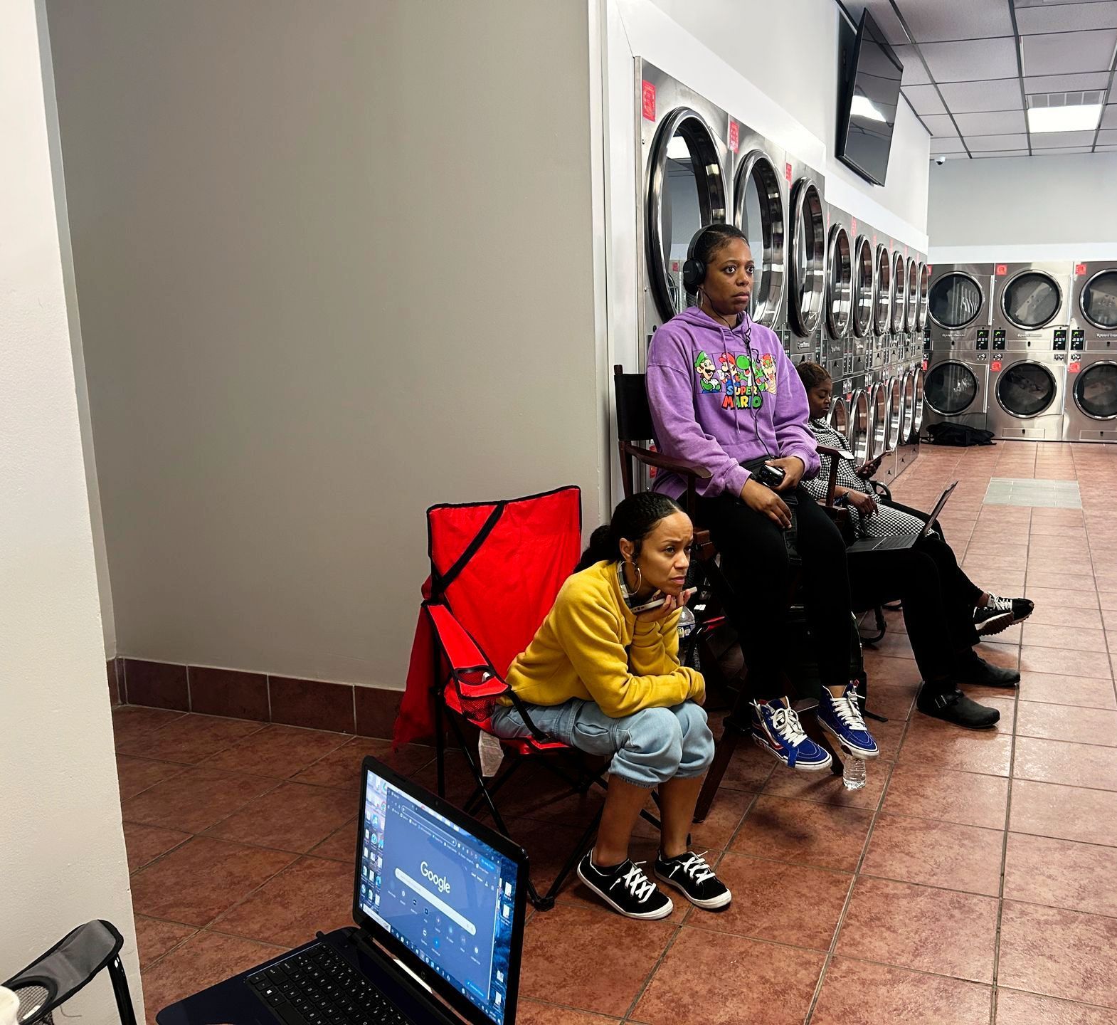 People waiting in a laundromat, sitting beside washers and a laptop on the floor.
