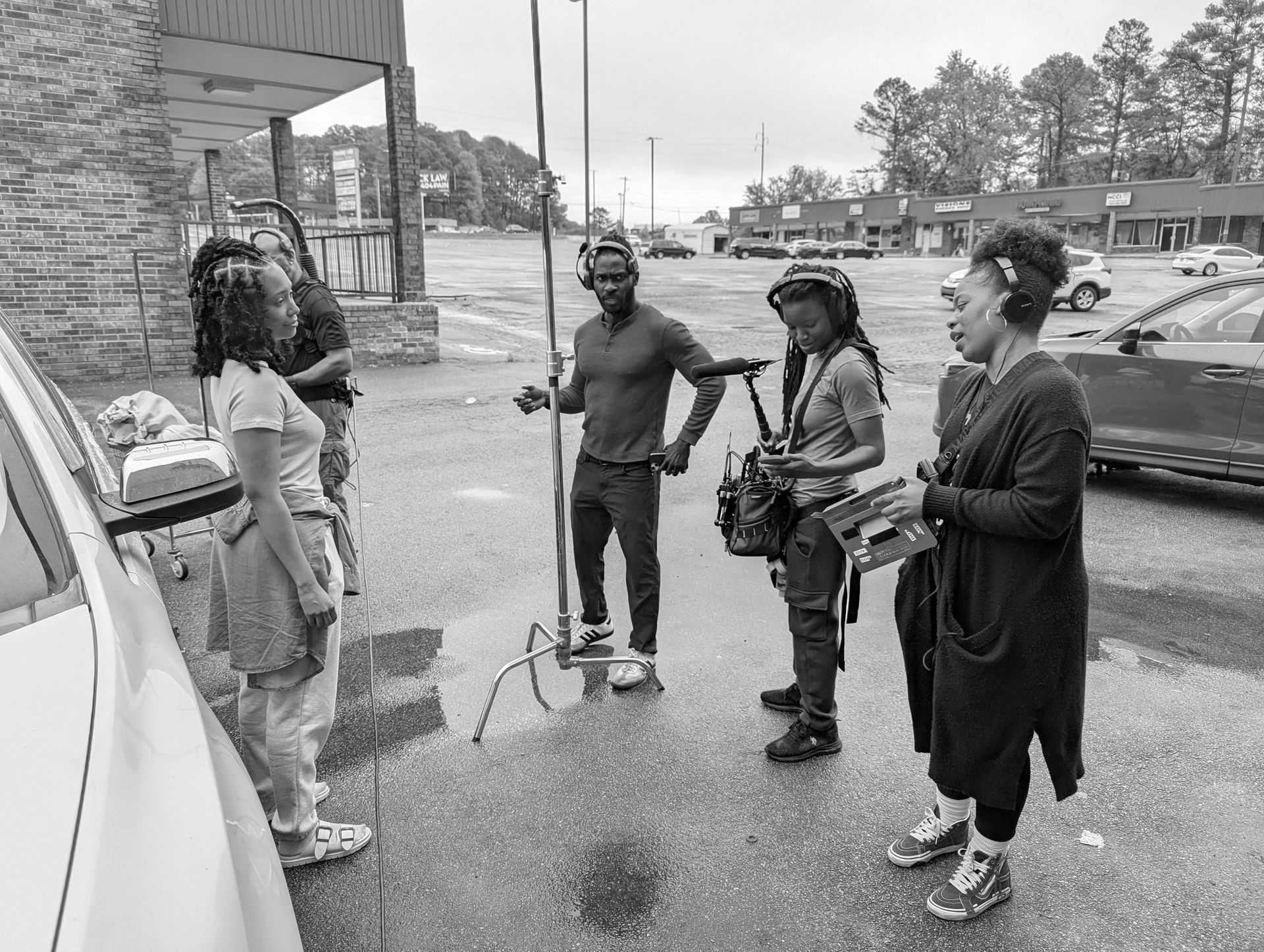 People gathered outside a store by parked cars, talking on a rainy sidewalk.