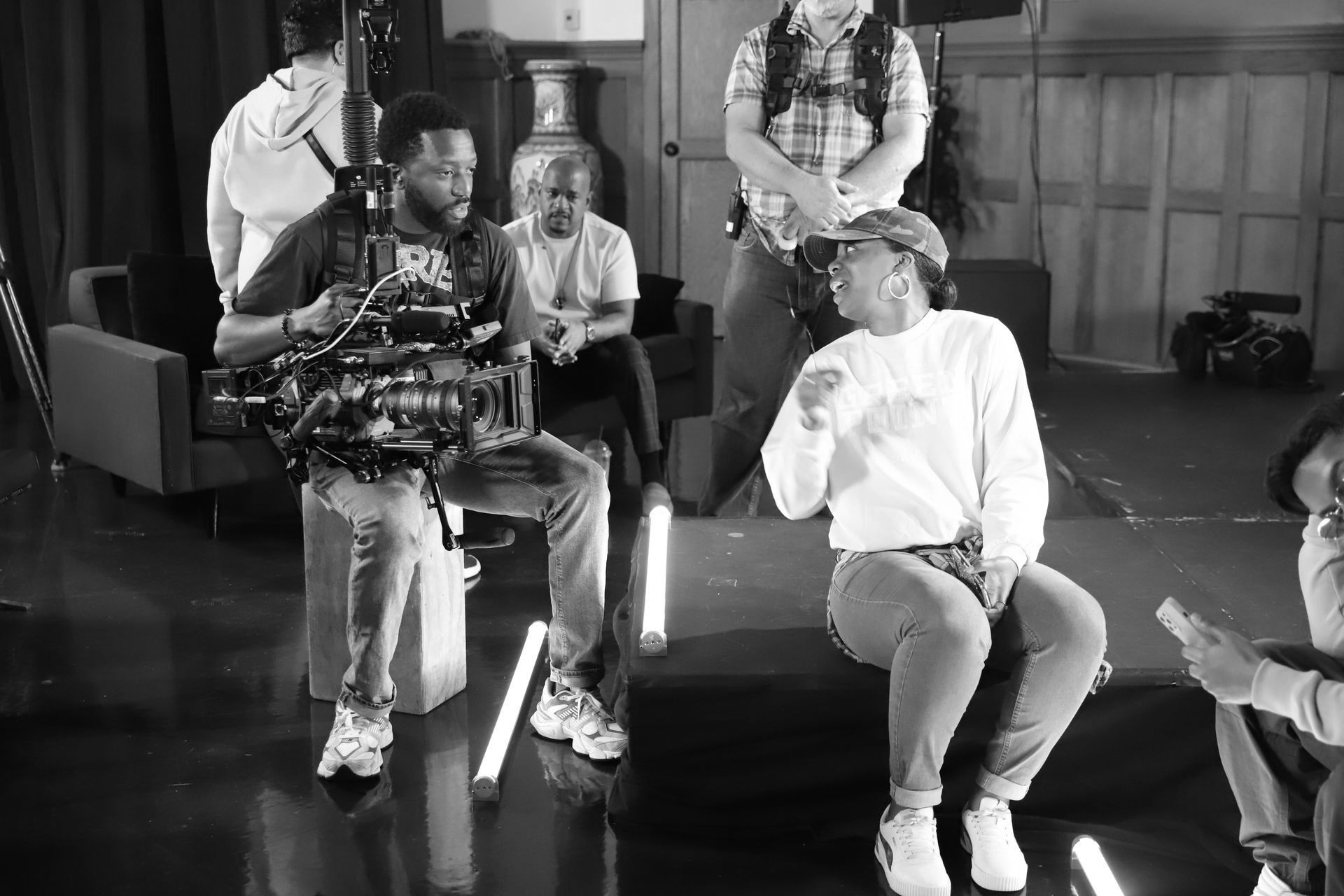 Two seated performers in a studio, one holding a guitar, with crew members standing behind them.
