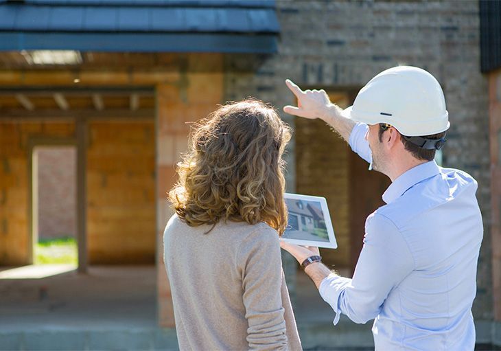 A man and a woman are looking at a tablet in front of a house under construction.