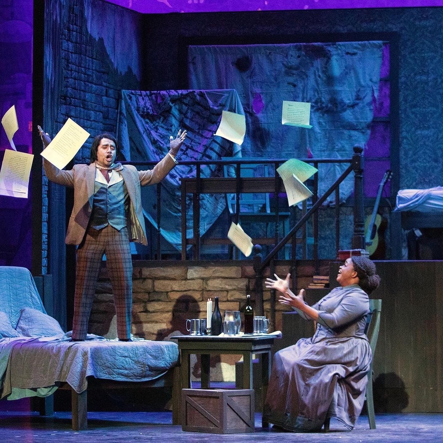 Two actors on a blue-lit stage with papers flying around a bed and small table.