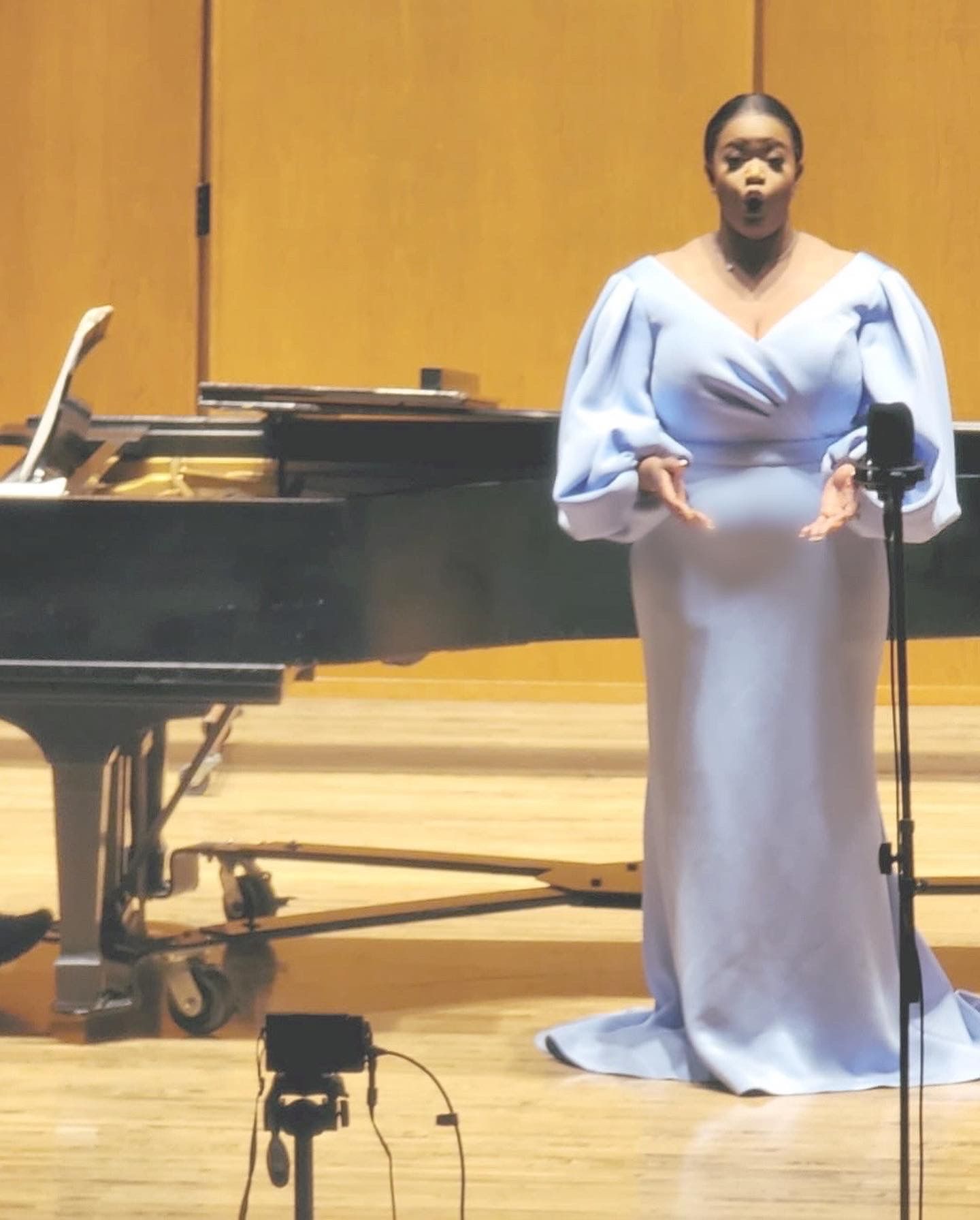 Singer in a light blue gown performing beside a grand piano on a concert stage