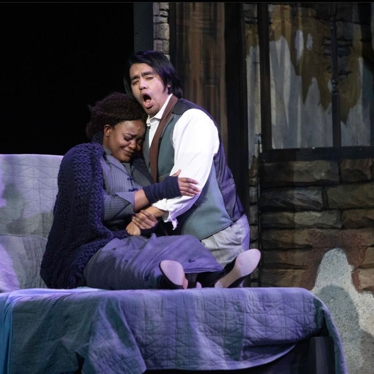 Two actors seated on a purple bed in a stage scene, one comforting the other, with rustic set pieces behind them
