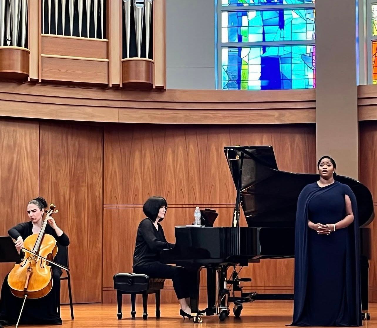Three musicians perform in a church: cello, piano, and a singer in a dark gown before wood paneling.