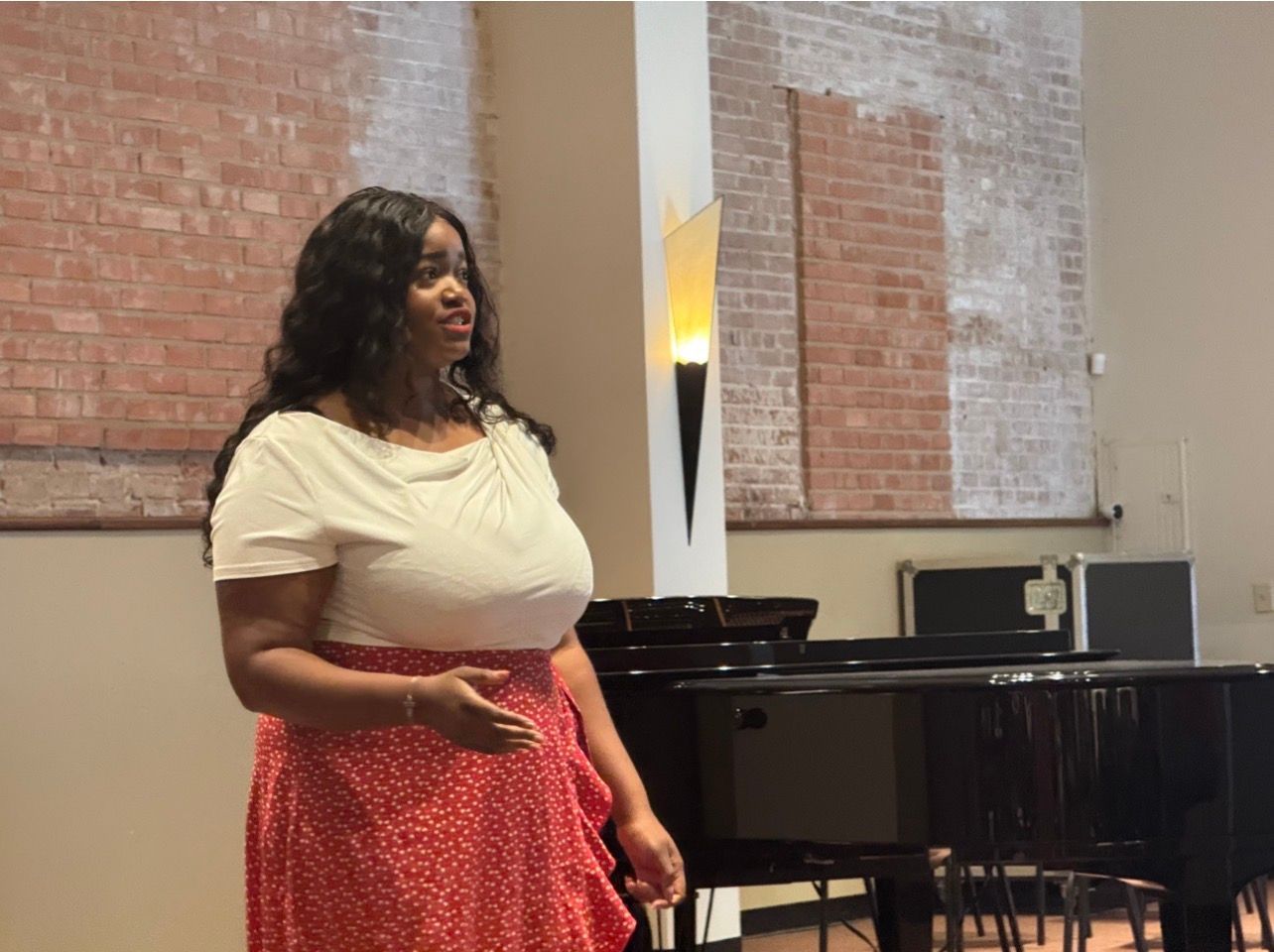 Person speaking in a room with a piano, wearing a cream top and red skirt.