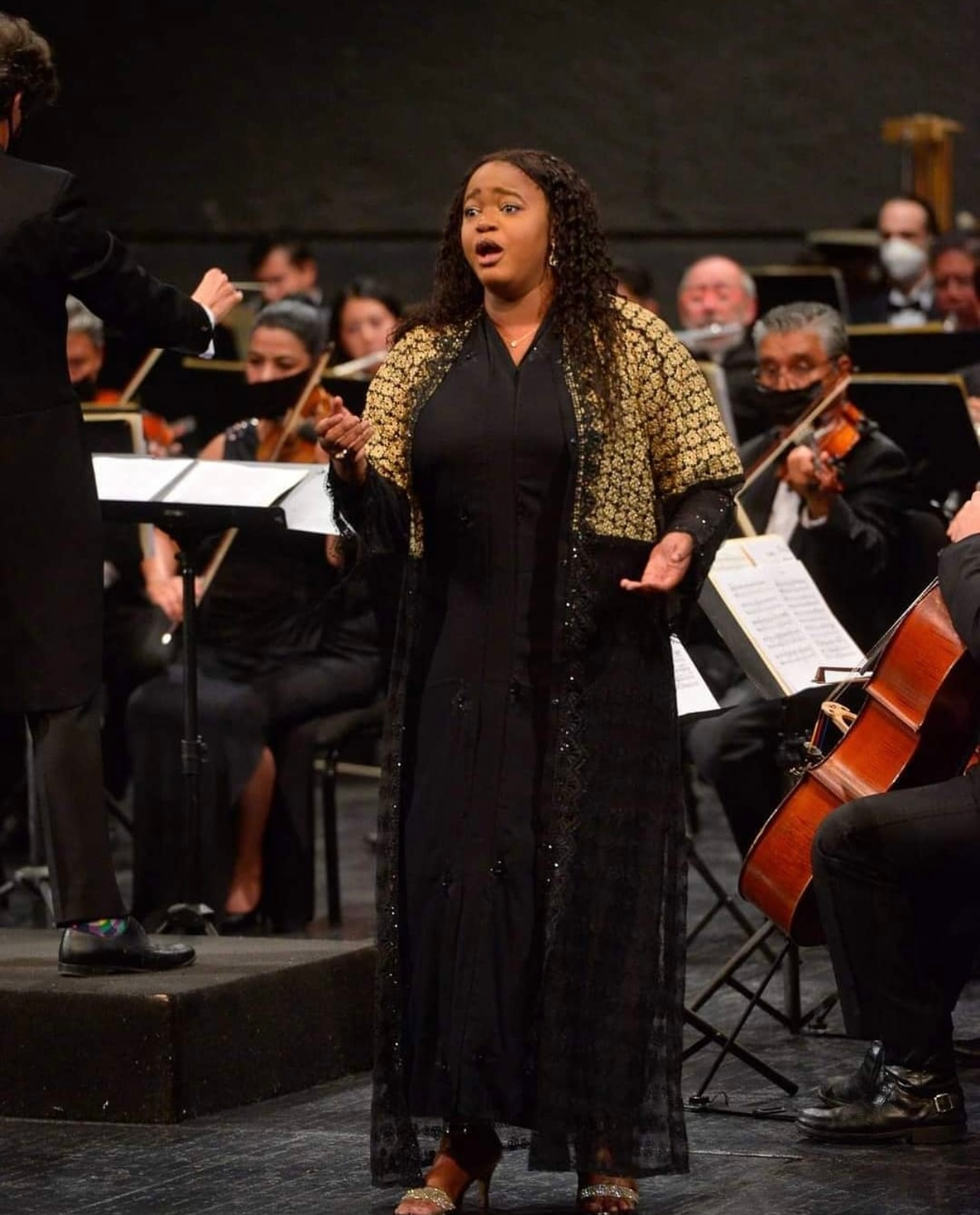 Opera singer performing onstage with orchestra, wearing a black gown and gold jacket, singing under stage lights.