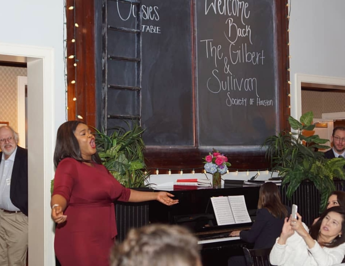 Woman singing at a piano in a decorated room with a chalkboard welcome sign and seated audience members