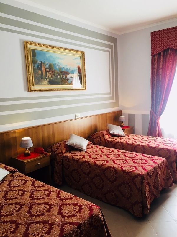 Tre letti in una stanza d'albergo con copriletti a fantasia rossa, opere d'arte e una finestra con tende rosse.