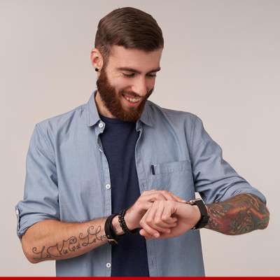 Homem barbudo sorrindo, olhando para o relógio no pulso. Vestindo camisa azul e camiseta escura. Tatuagens nos braços.