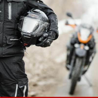 Motociclista com equipamento preto segurando capacete, motocicleta ao fundo.