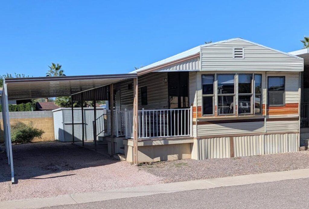Single-story mobile home with a carport and porch under a bright blue sky.