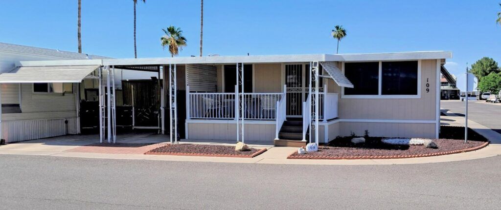 White mobile home with a front porch and carport in a desert neighborhood under a blue sky