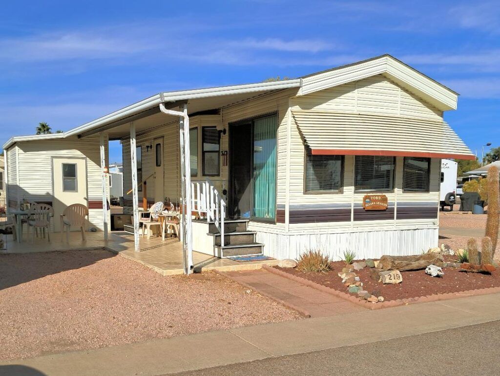Small white manufactured home with a covered porch and gravel yard in a desert setting