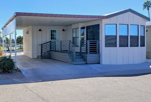 Single-story beige mobile home with a front ramp and steps, shown on a sunny street corner.
