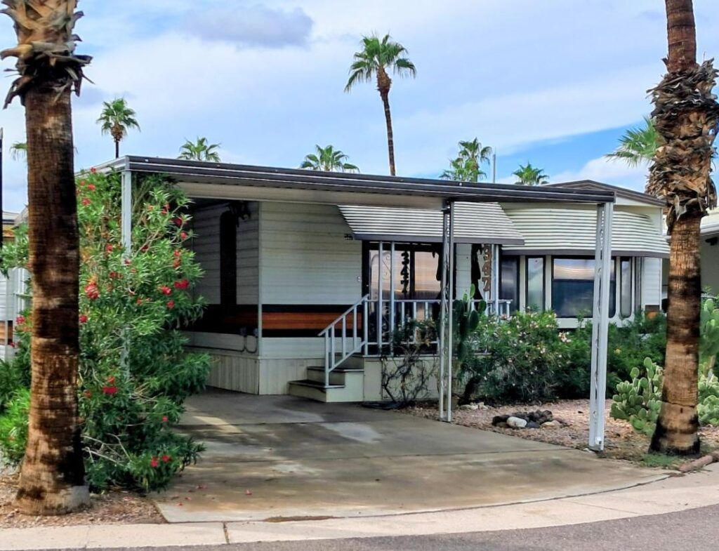 Mobile home with porch and steps, framed by palm trees and shrubs in a sunny driveway.