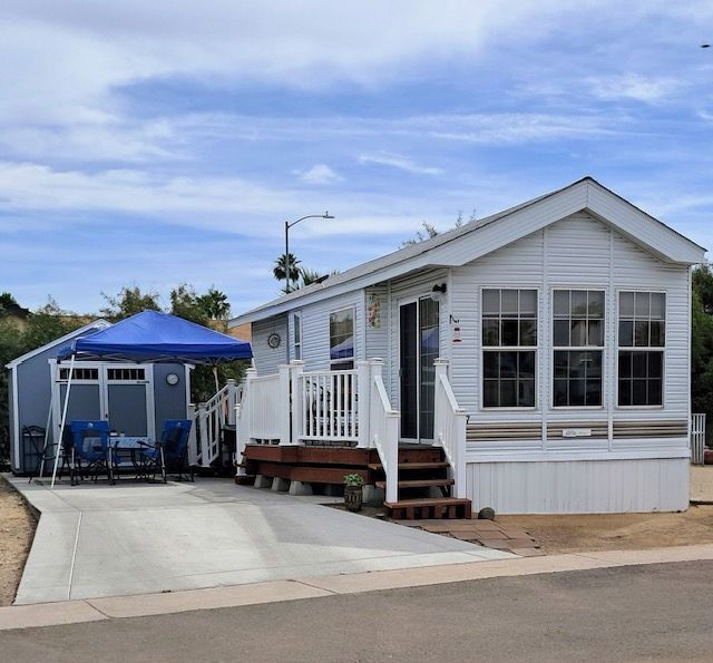 White manufactured home with porch and blue canopy beside a concrete driveway under a cloudy sky