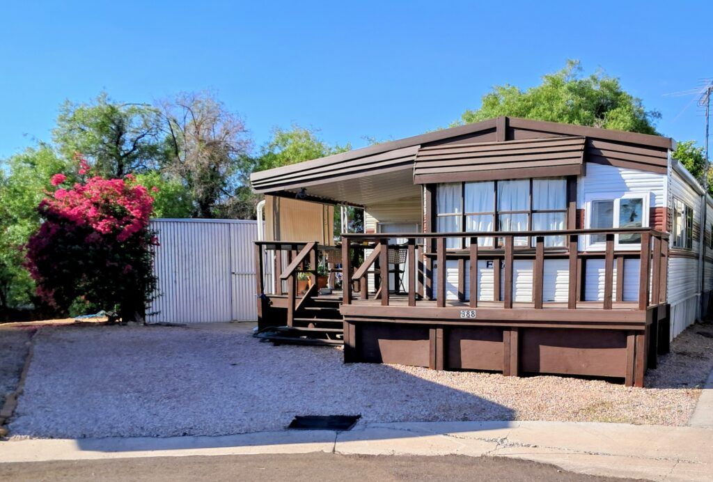 Modular home with a wooden deck, gravel yard, and a pink flowering bush under a blue sky