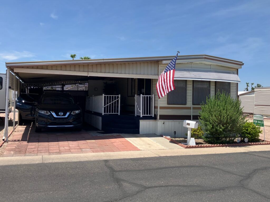 Single-story mobile home with a carport, parked SUV, and an American flag in front under a clear sky