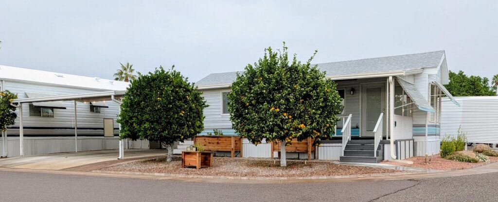 White mobile home with two trees and a bench in front on a gravel lot