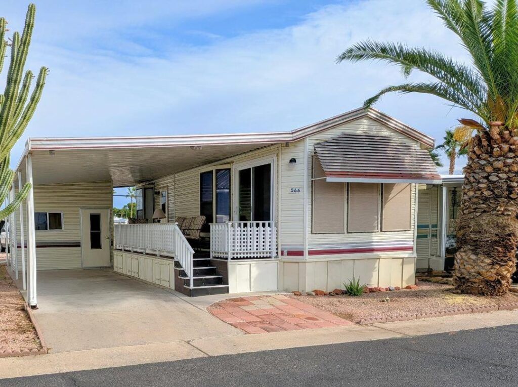 Mobile home with a front porch, white trim, and a palm tree beside it under a clear sky.