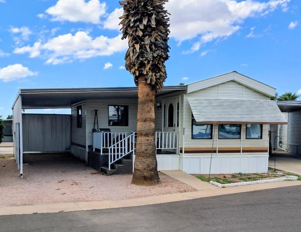 Mobile home with a carport and a palm tree in front, on a quiet residential street.