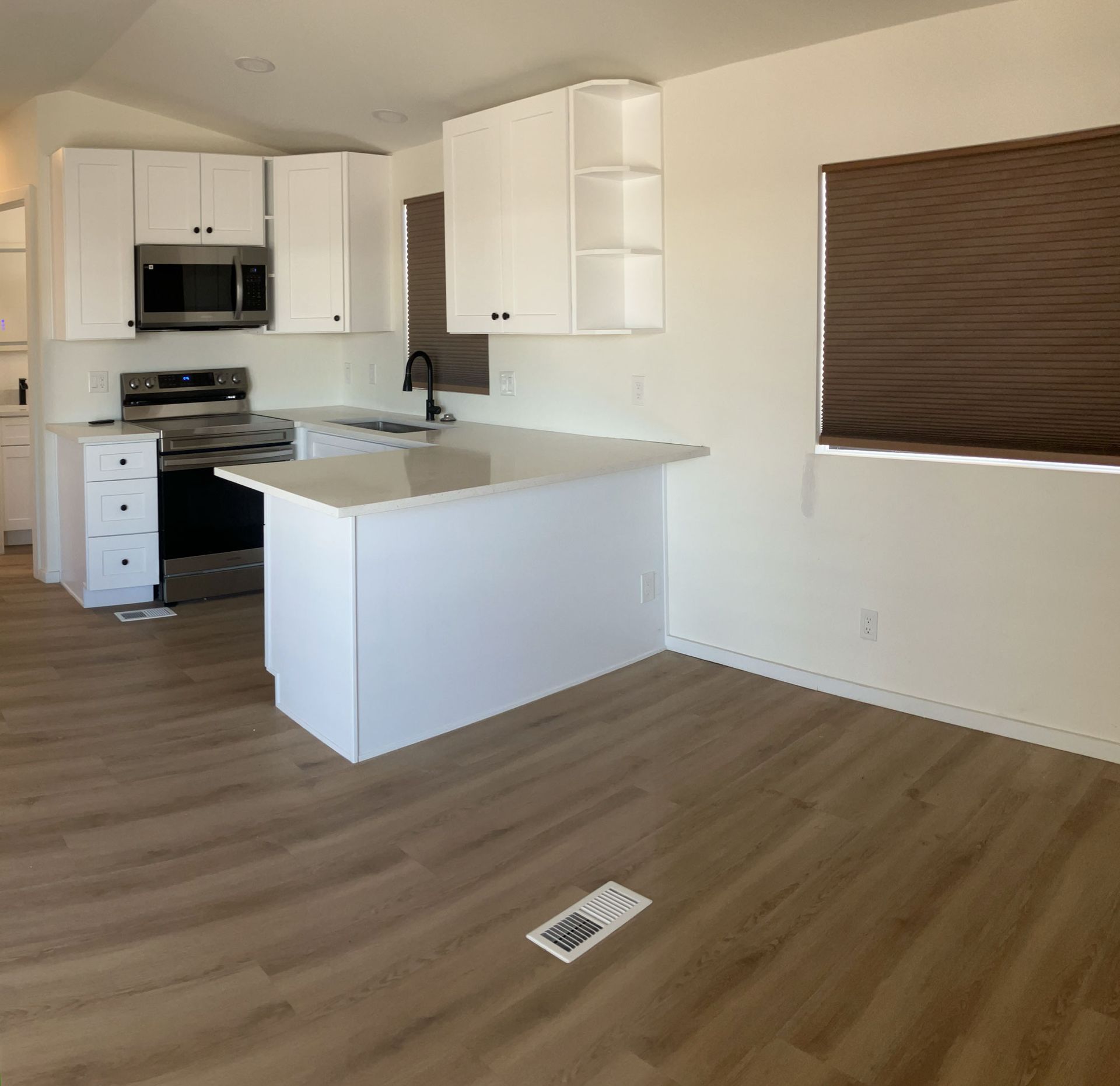 Empty modern kitchen and living area with white cabinets, island, and wood floors, showing a window with blinds.