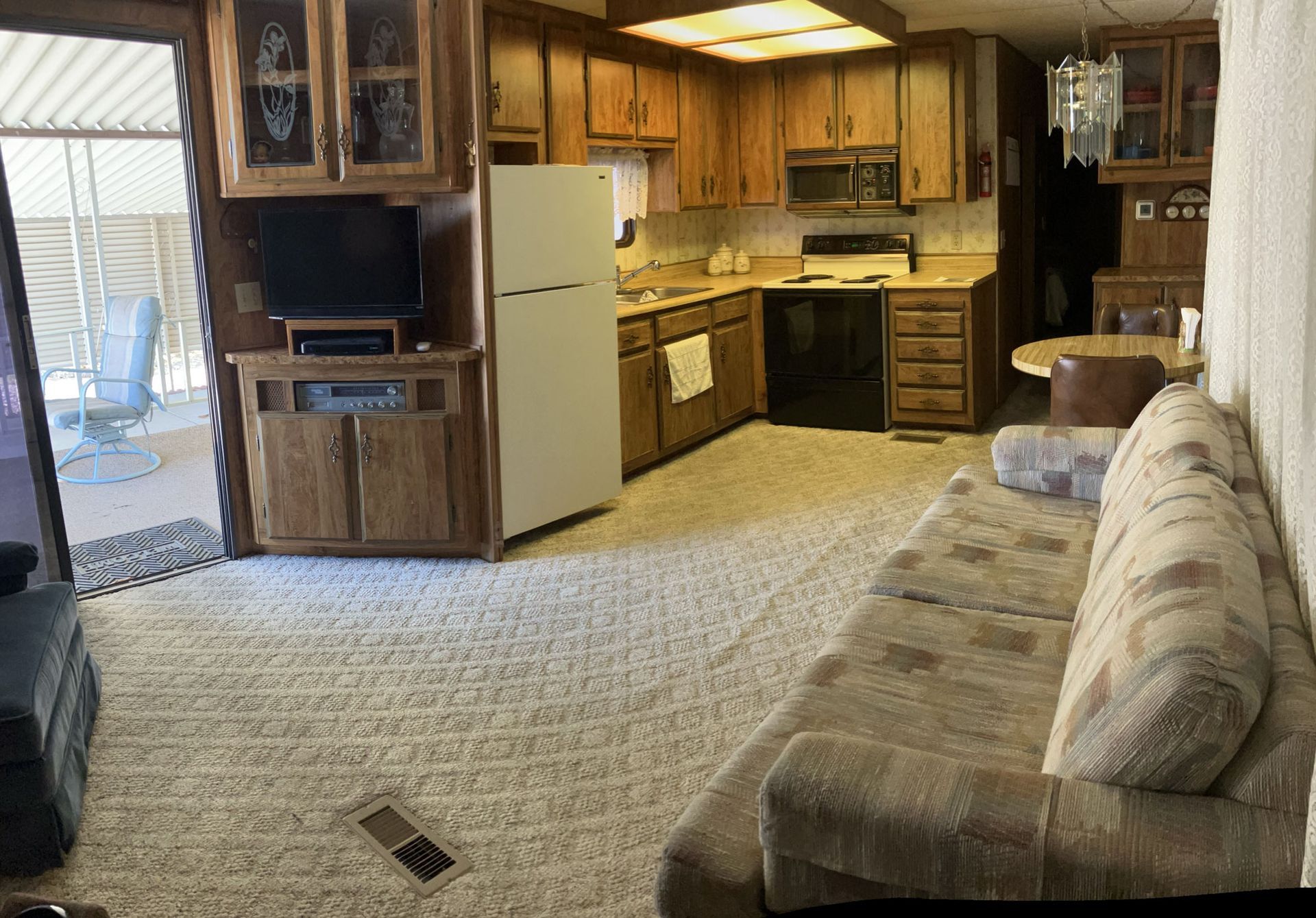 Small living room with wood cabinets, beige sofa, carpet, and kitchen area in the background.