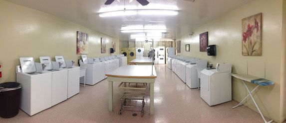 Bright laundry room with rows of washing machines, folding tables, and a central cart under fluorescent lights