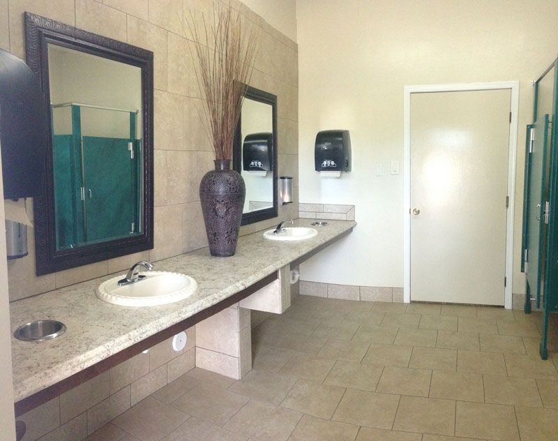 Public restroom with long granite sink counter, mirrors, vases, beige tile floor, and closed door