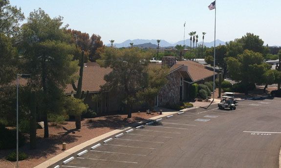 Desert-style parking lot beside low buildings and trees, with a flagpole and mountains in the distance