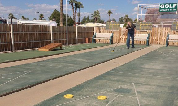 Outdoor mini-golf course with green lanes, wooden fences, and two people near the far end under a sunny sky
